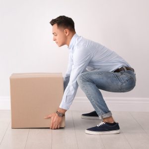 Full length portrait of young man lifting heavy cardboard box near white wall. Posture concept